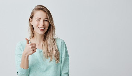 Happy young caucasian female wearing blue long sleeved shirt making thumb up sign and smiling cheerfully, showing her support and respect to someone. Body language. I like that. Good job.