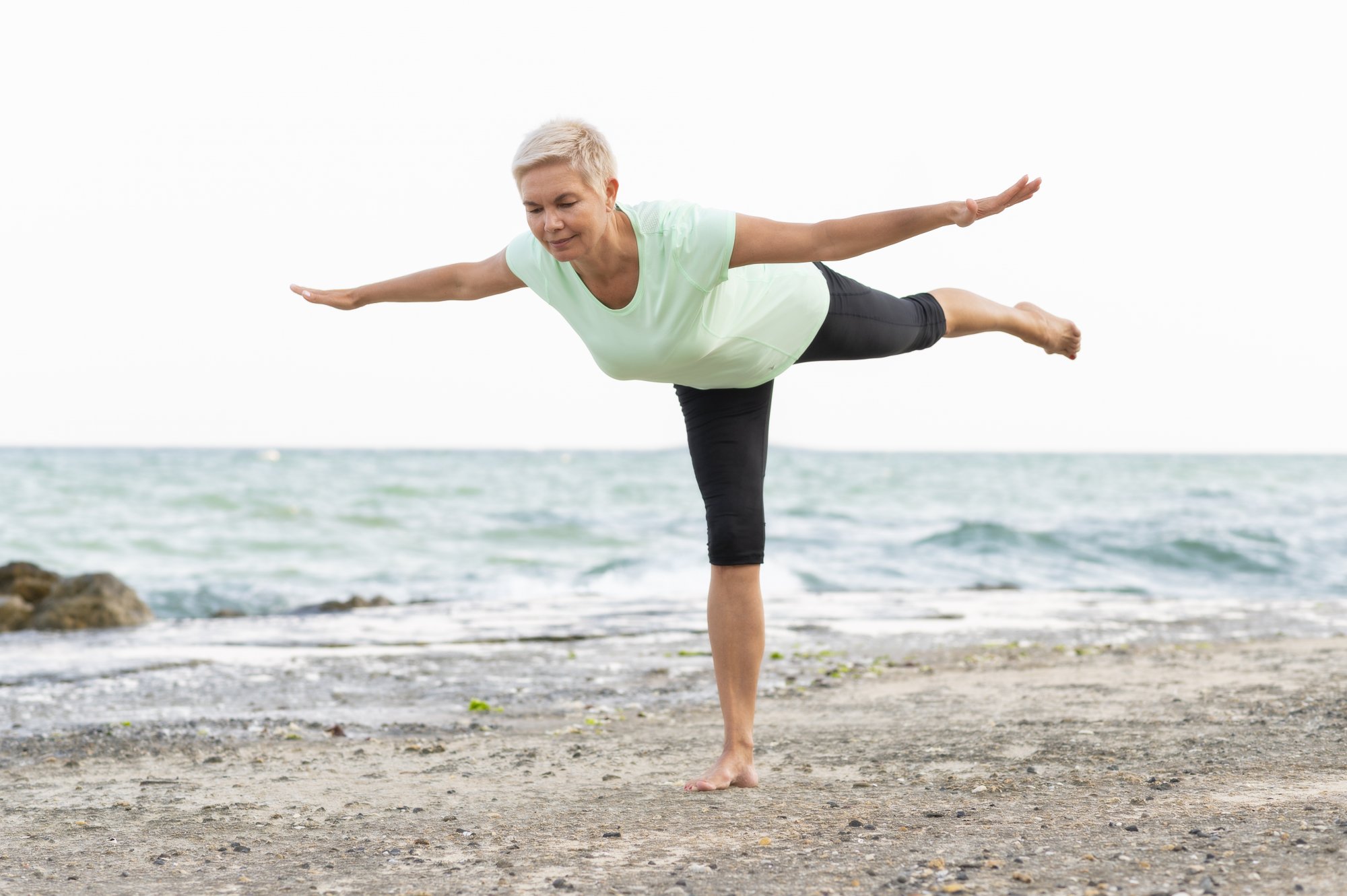 Balancetraining2.jpg Eine Frau in einem hellgrünen T-Shirt und schwarzen Leggings balanciert auf einem Bein am Strand.