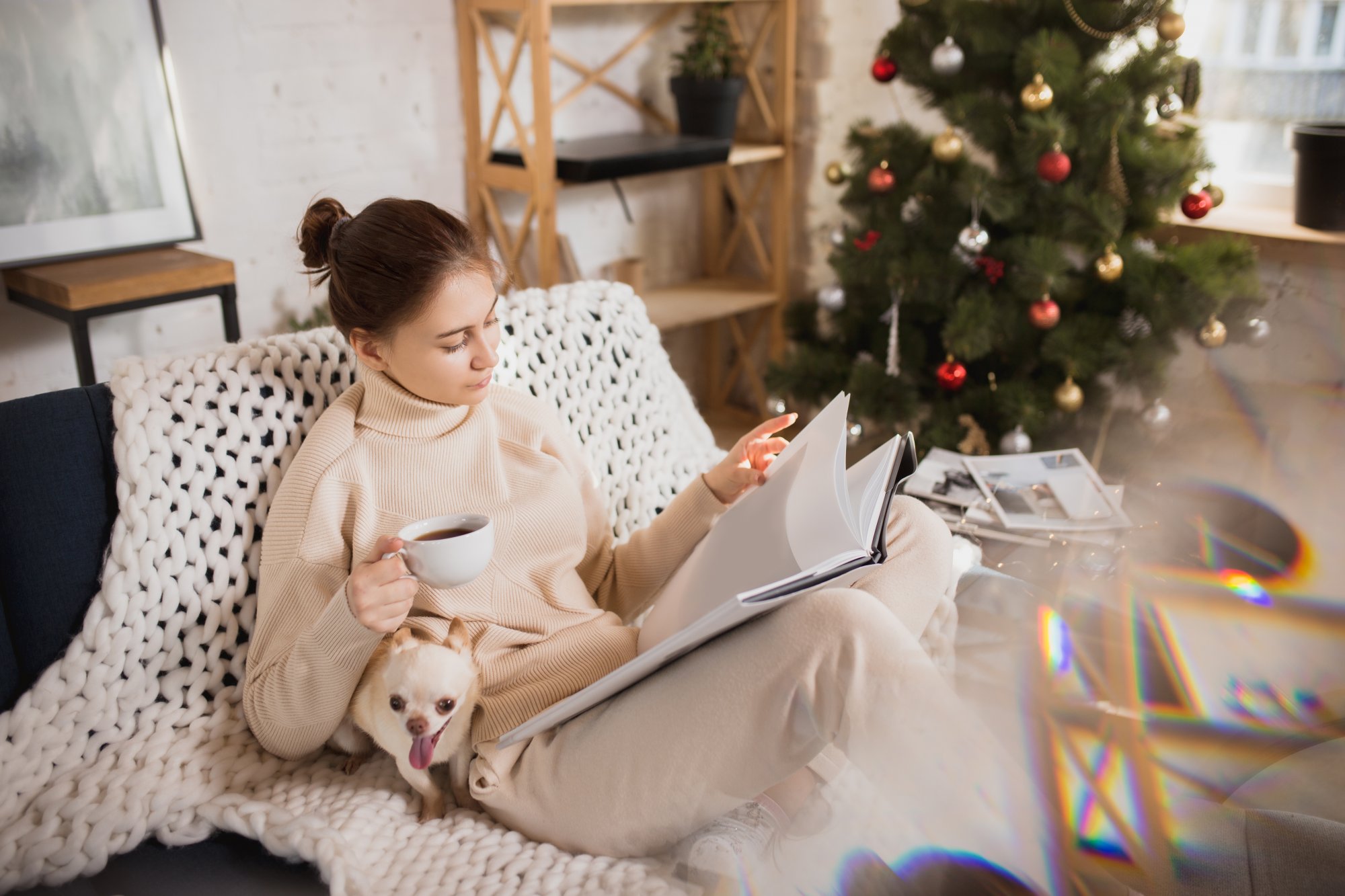 Immer_mit_der_Ruhe.jpg Eine Frau in bequemer Kleidung sitzt auf einem Sofa, liest in einem Buch und hält eine Tasse Kaffee, während ein Hund bei ihr sitzt.