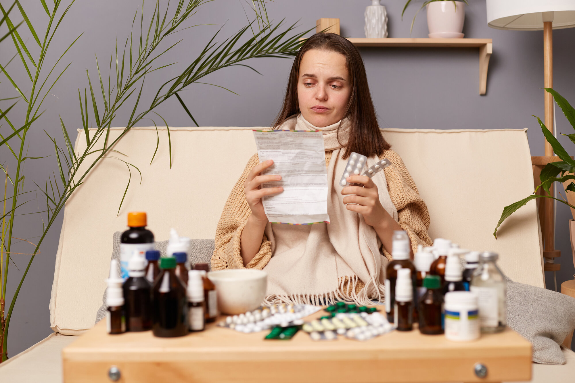 Eine kranke Frau sitzt auf einem Sofa, umgeben von verschiedenen Medikamenten und hält einen Zettel sowie eine Medikamentenpackung in der Hand.